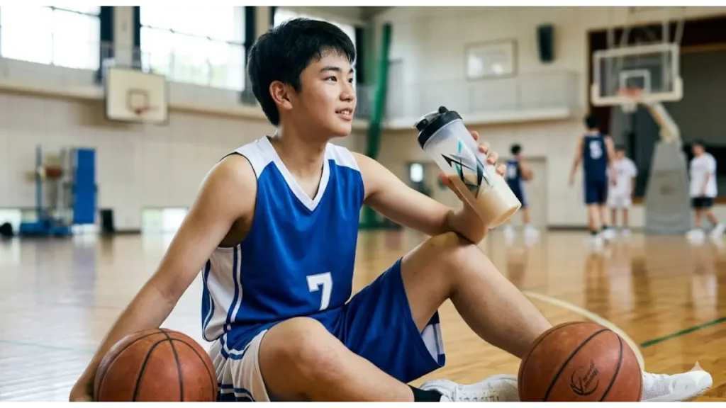 体育館の床に座り、練習後に栄養シェイク(ノビエースを想定した translucent shaker bottle)を飲む日本人の男子中学生バスケ部員。体格づくりへの意識を表現(実写風)。