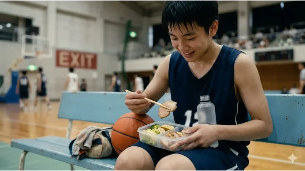 試合後の体育館ベンチで、疲労回復のために鶏チャーシューや卵焼きなどの高タンパク質おかずが入った弁当を食べる日本の男子中高生バスケットボール選手。