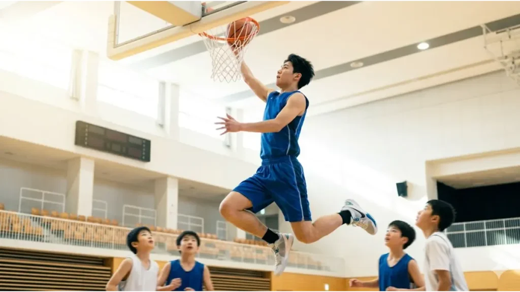 305cmのバスケットゴールに向かってジャンプするアジア人の少年と、それを下から見上げる身長の低い選手たち。